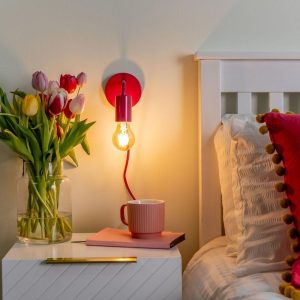 Plug in wall light in raspberry red, shown lit on bedroom wall